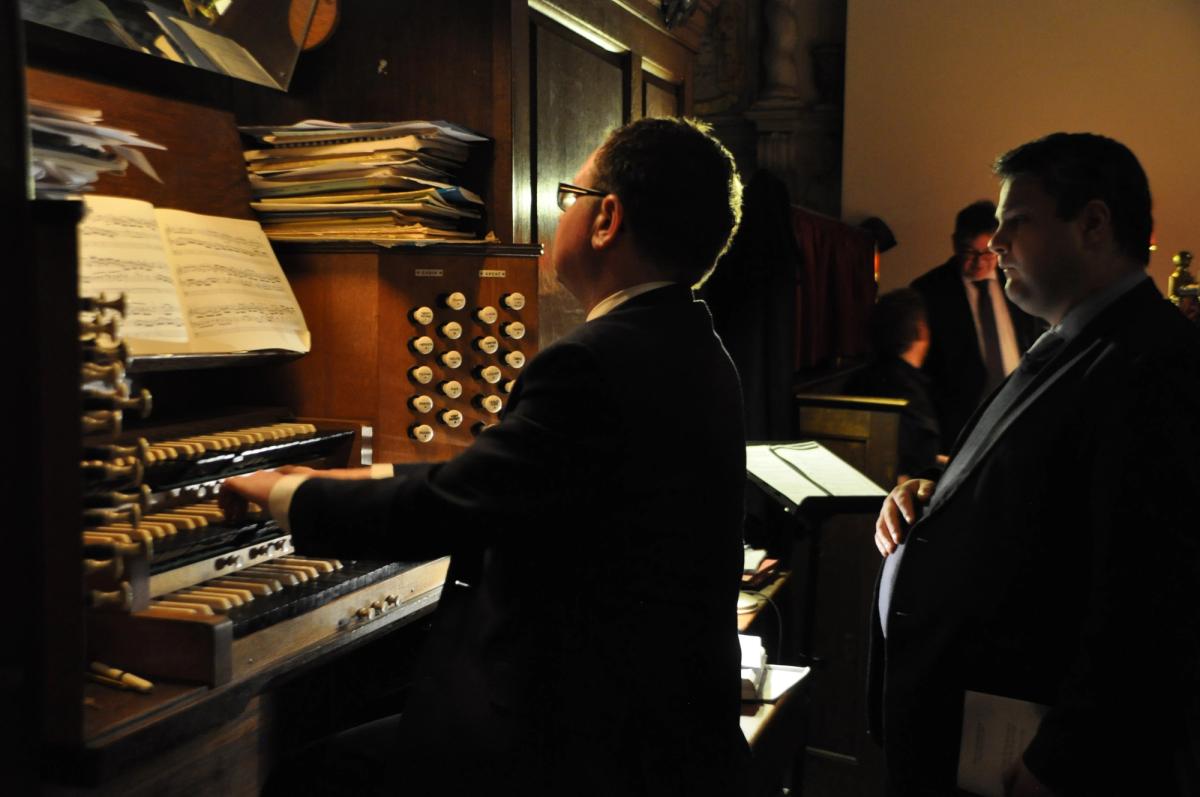 An organist playing the organ while another person stands alongside.