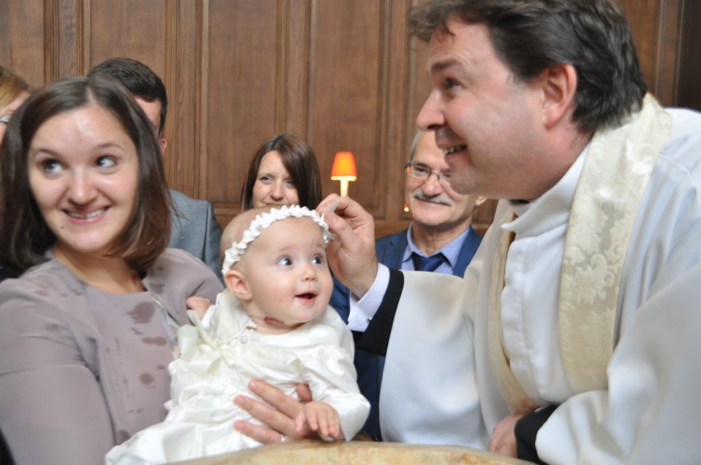 A baby in white held beside a priest and a lit candle.
