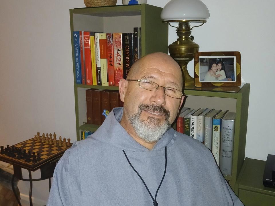 A seated man in grey clothing wearing a large cross pendant.