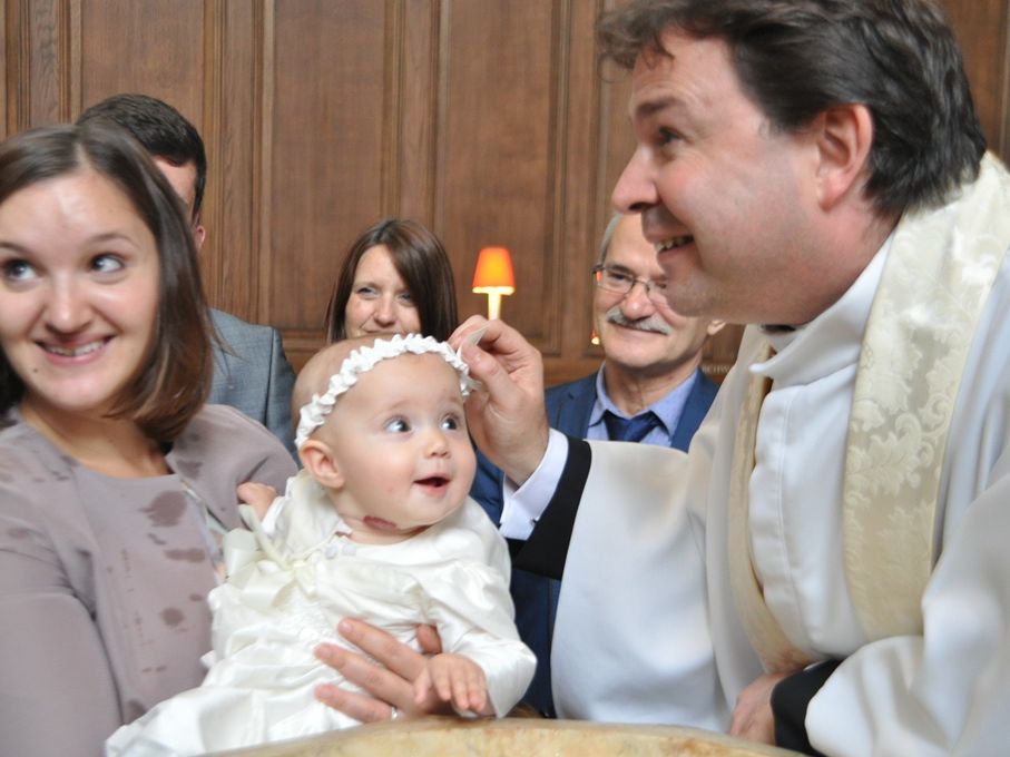 A baby in white held beside a priest and a lit candle.