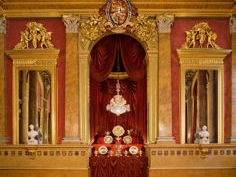 An ornate gilded ceremonial room with a red chair and decorative panelling.