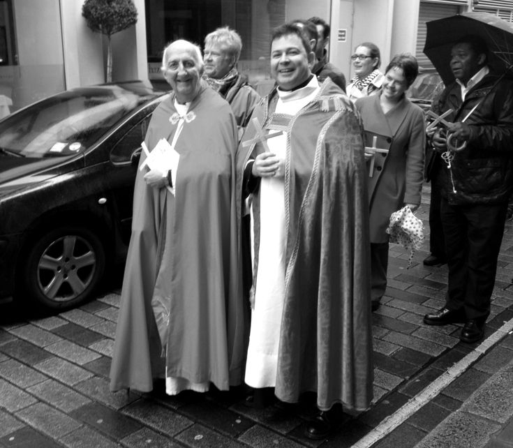 Three members of the clergy standing on the pavement with worshippers behind.