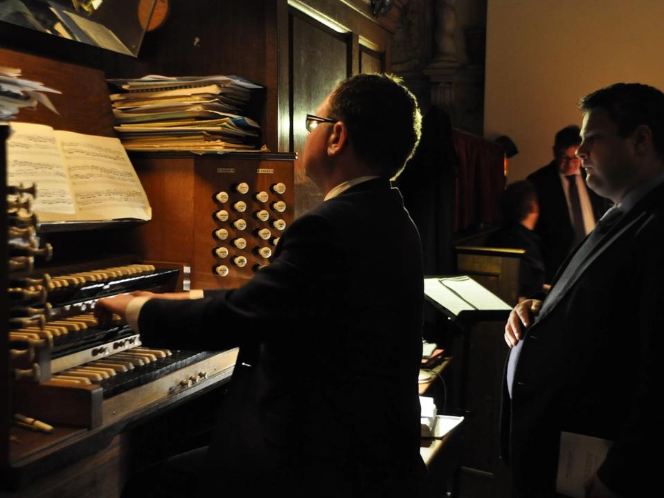 An organist playing the organ while another person stands alongside.